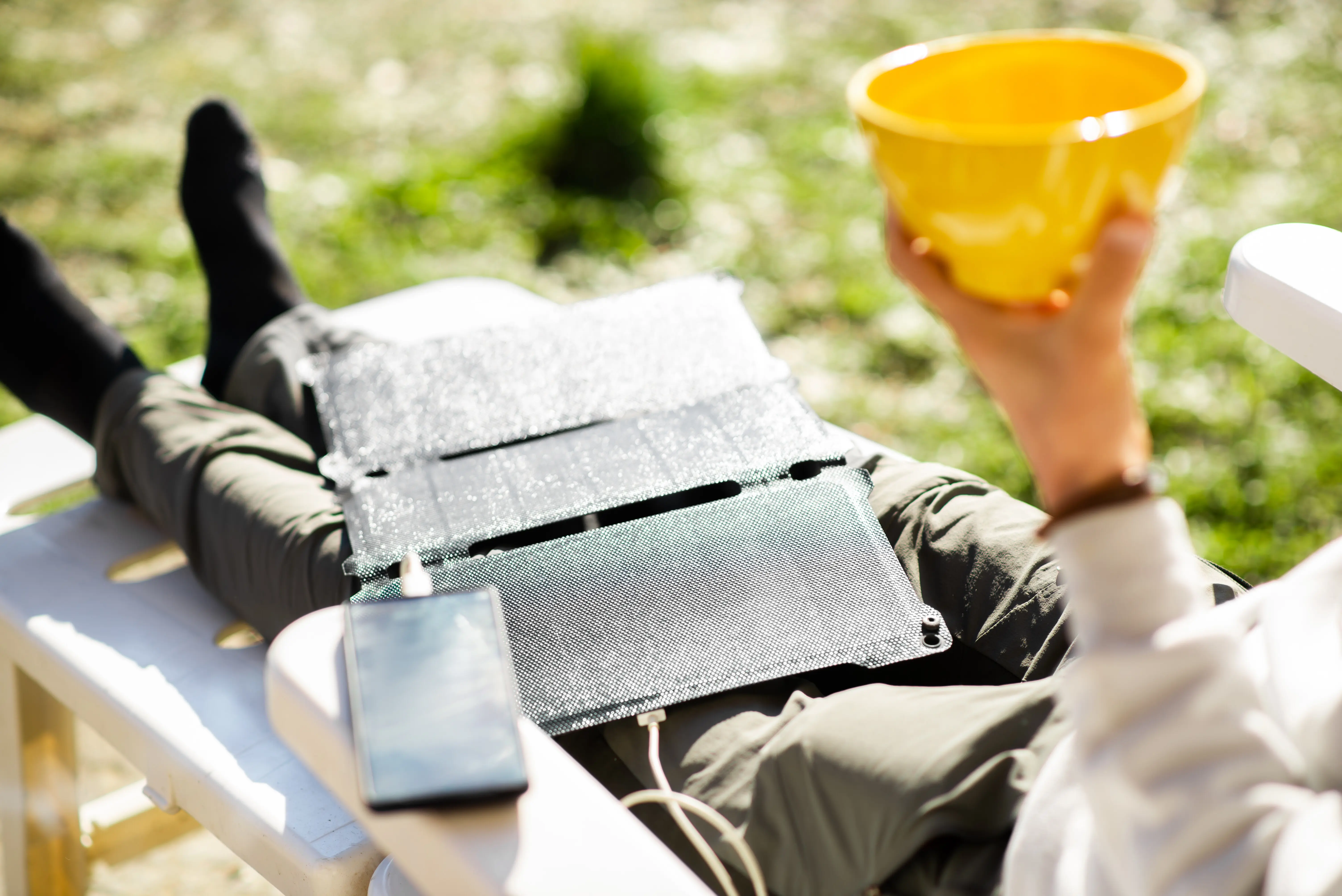 Auf den Beinen eines ruhenden, nicht erkennbaren Mannes liegt eine tragbare Solarbatterie, die ein Mobiltelefon lädt – ökologische Energie.